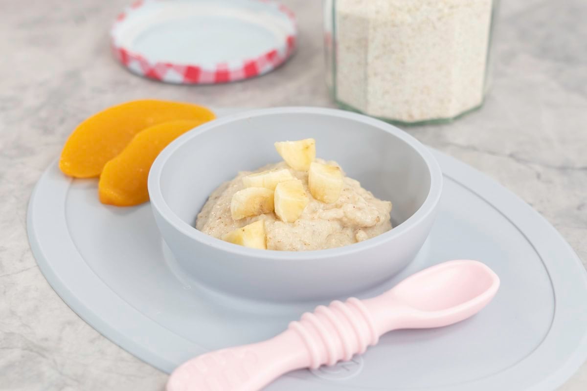 Silicone baby bowl and placemat on marble bench top with baby oats inside, topped with chopped banana pieces and two slices of canned peaches on the side. Pink baby spoon and jar of ground oats on benchtop.