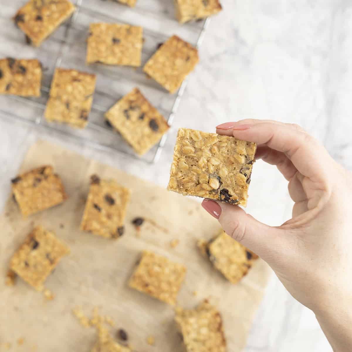 Woman's hand holding a piece of oat slice above a cooling tray and baking paper on bench top with slice on top.