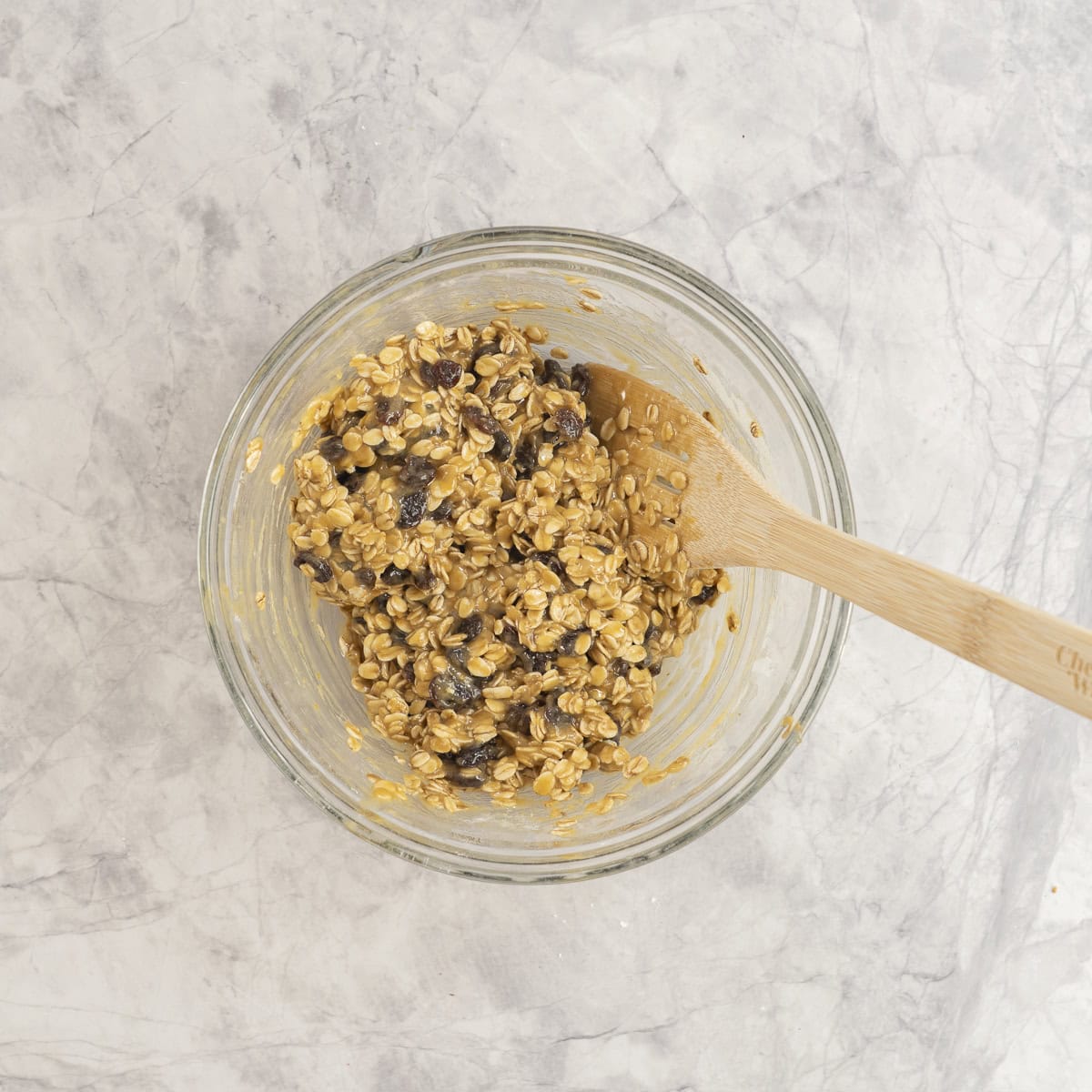 Oats and raisins added into mixture inside glass bowl with wooden spoon on marble bench top.