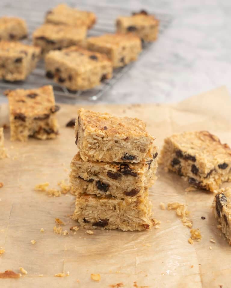 Three pieces of cooked oat slice staked on baking paper on benchtop, sliced pieces in background on cooling tray.