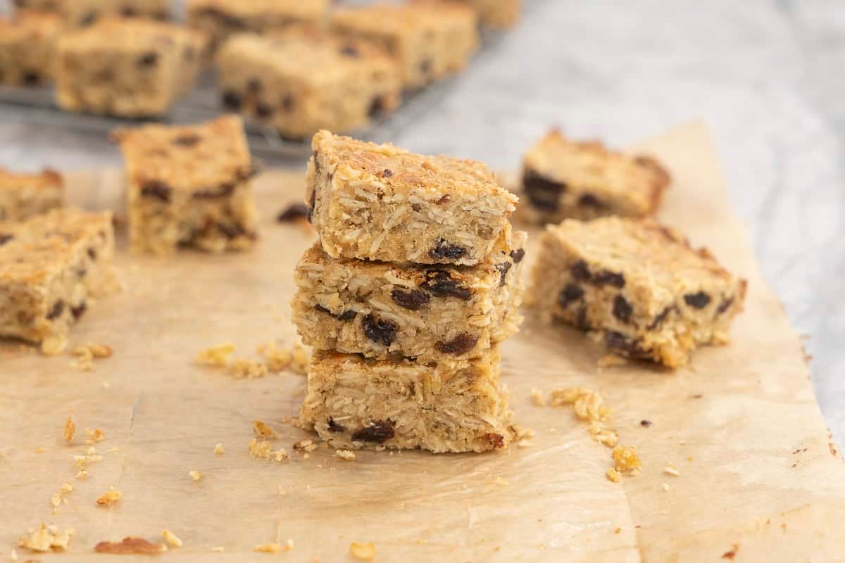 Three pieces of cooked oat slice staked on baking paper on benchtop, sliced pieces in background on cooling tray.