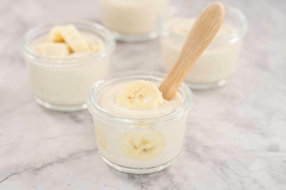 Four small glass ramekins with banana yogurt and fresh slices of banana inside, one with a toddler spoon.