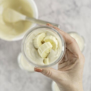 Woman's hand holding up a small glass ramekin with yogurt and banana slices inside, more filled ramekins below on bench top and white bowl with spatula inside.