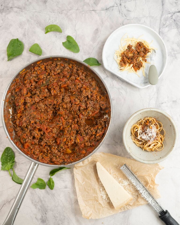 Pan on marble benchtop with bolognese mince inside, a dinner plate and bowl served with spaghetti pasta, spinach leaves scattered on bench top.