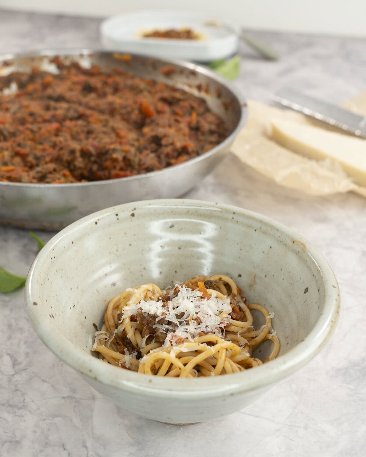 Dinner bowl with a serving of spaghetti with bolognese in a sauce pan in background on benchtop.