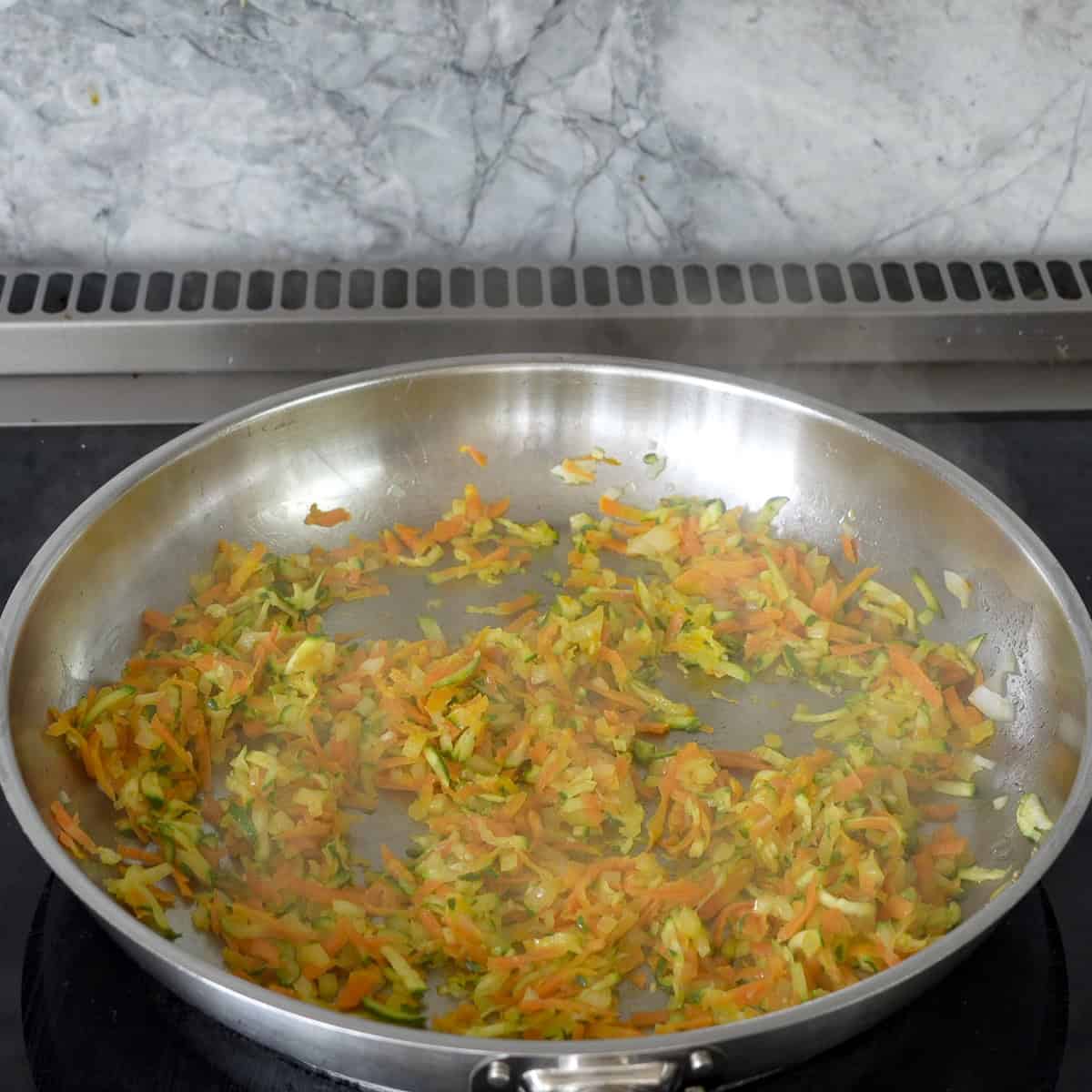 Grated carrot, zucchini, and onion in a large pan on stove top.