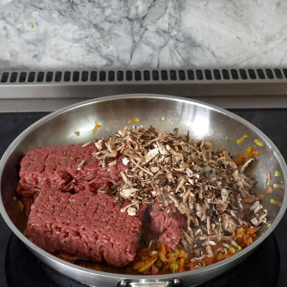 Grated mushroom and mince added into cooked grated carrot, zucchini, and onion in a large pan on stove top.
