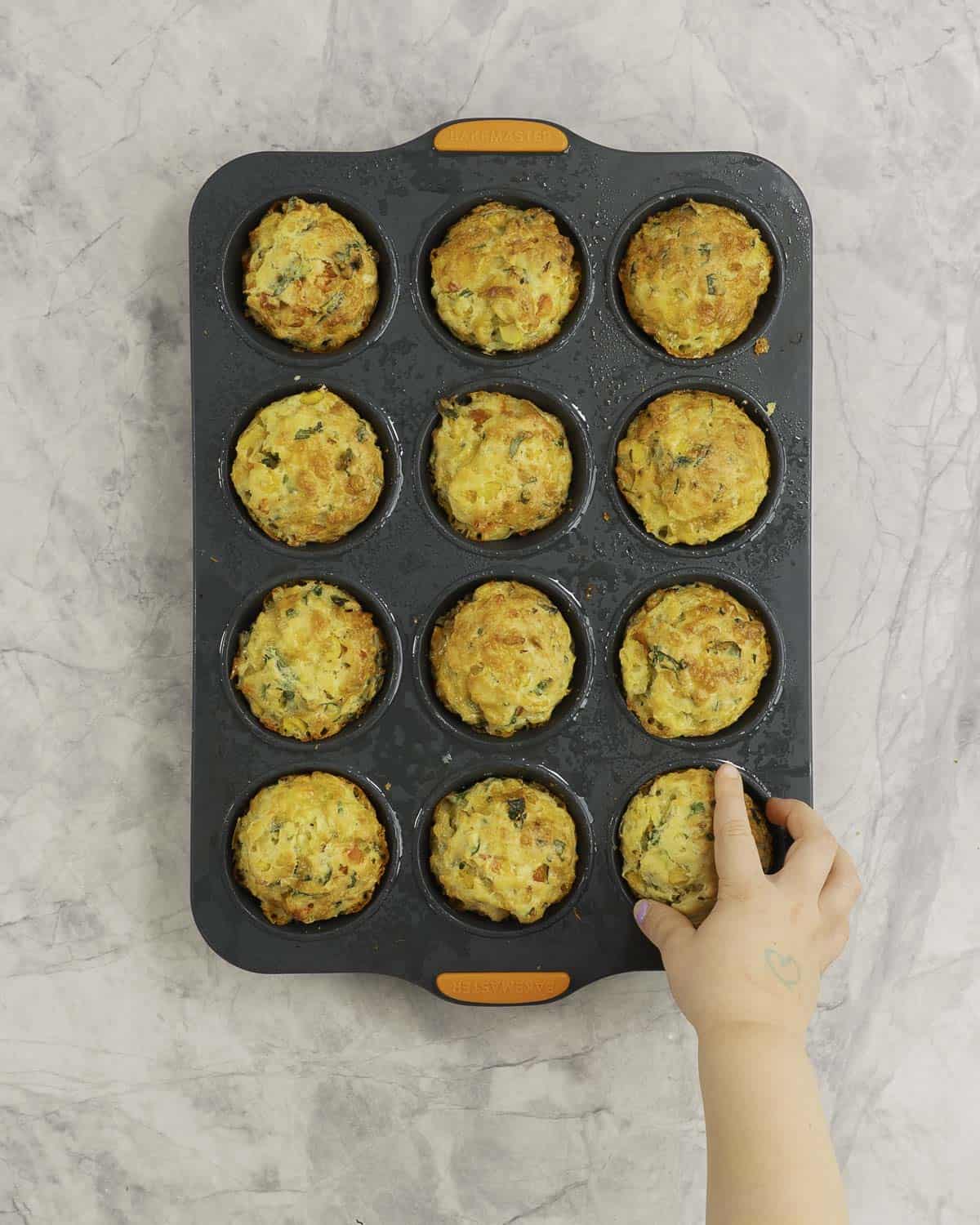 Toddlers hand reaching to take a muffin from a muffin tray on bench top.