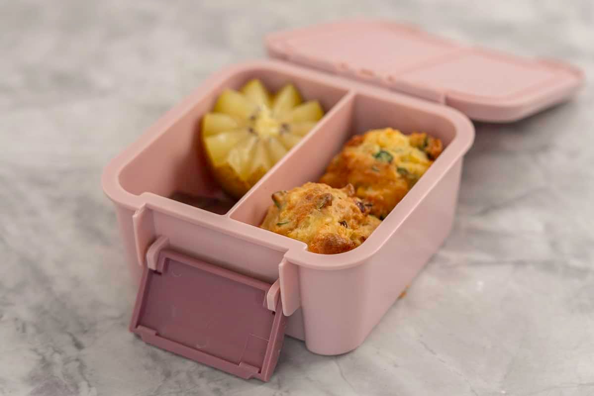 Small pink lunch box with two vegetable muffins in one compartment and a halved kiwifruit and chocolate slice.
