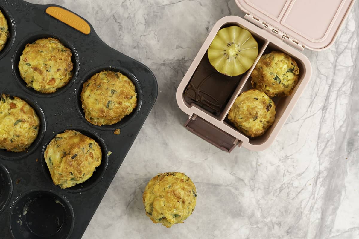 Small pink lunch box with two vegetable muffins in one compartment and a halved kiwifruit and chocolate slice, muffin tray with more inside on benchtop.
