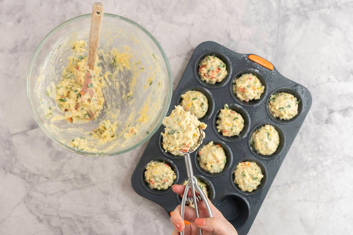 Cookie scoop with vegetable muffin batter inside serving into a muffin tray on bench top with empty glass bowl.