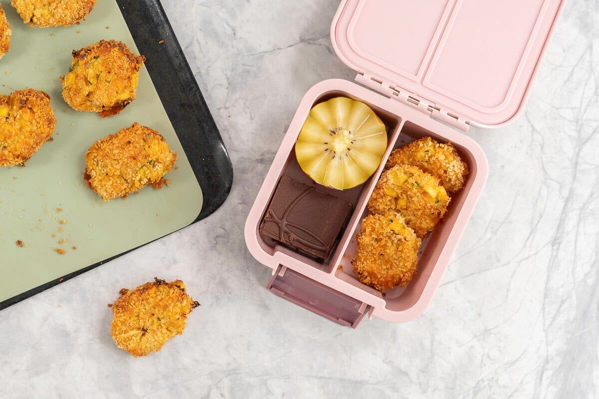 Lined baking tray with crispy nuggets on bench top and a small pink lunch box with three nuggets inside, a halved kiwifruit and chocolate slice.