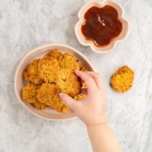 Toddlers hand taking a nugget from a stack on a plate, tomato sauce in ramekin and nugget on marble bench top.