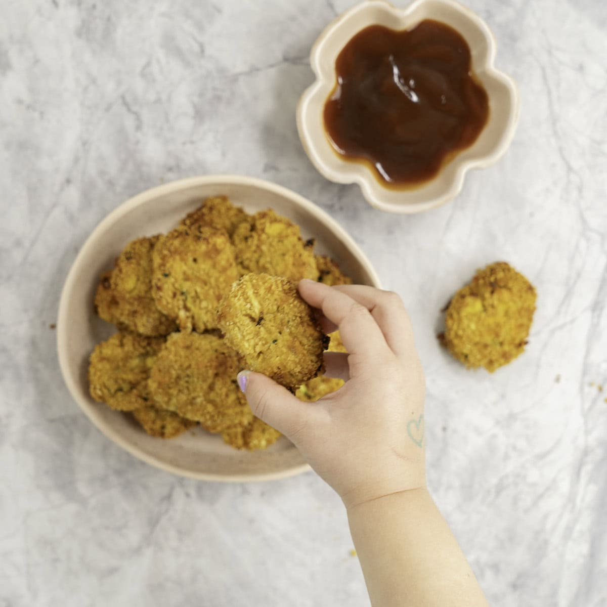 Toddlers hand taking a nugget from a stack on a plate, tomato sauce in ramekin and nugget on marble bench top.