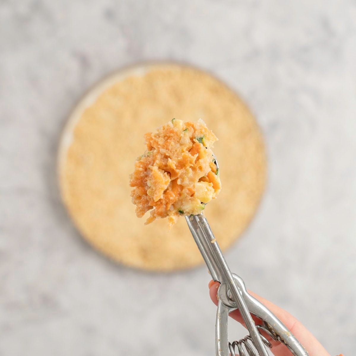 Cookie scooper with a scoop of vegetable mix inside help up to camera with a plate of crumbs on marble bench top below.