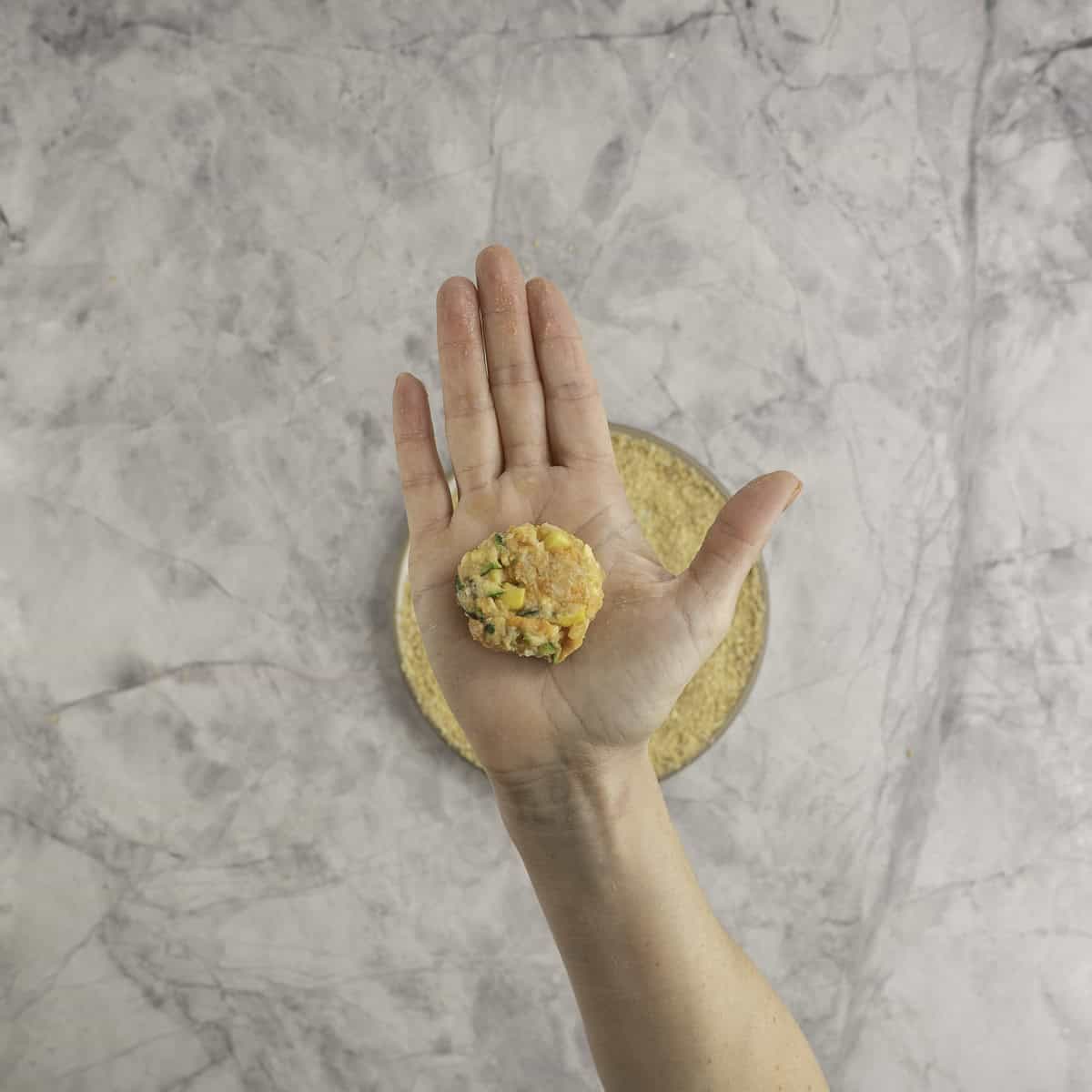 A small circle nugget in the palm of a woman's hand help up to camera with a plate of crumbs on marble bench top below.