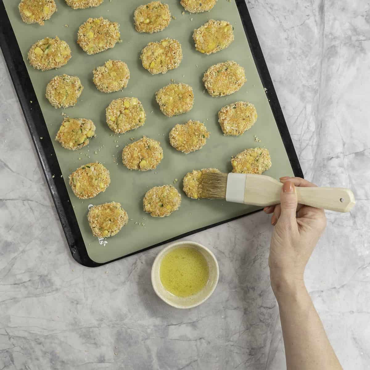 Lined tray on marble benchtop with small oval shaped nuggets crumbed, hand brushing melted butter on top.