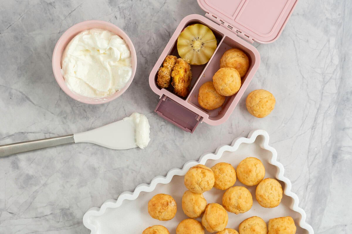 White rectangle dish with wavy border, batch of mini muffins inside, small pink ramekin with greek yogurt inside and a spatula on bench top, a small pink lunch box with three muffins inside, halved kiwifruit and homemade nuggets.