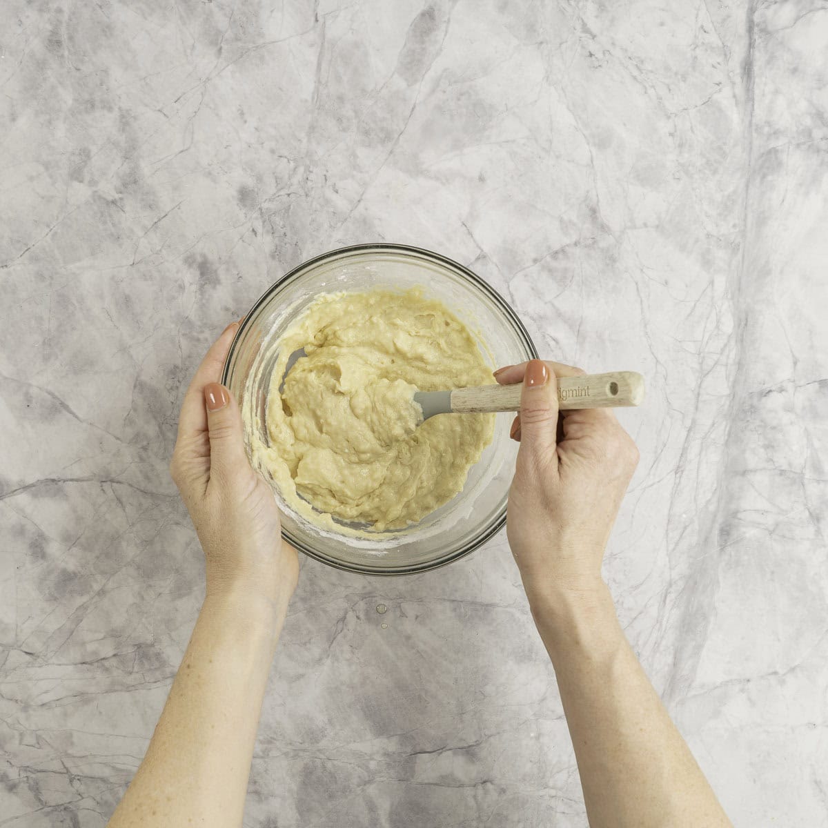 A small glass bowl on marble bench top with wet ingredients being stirred with a spatula by a woman.