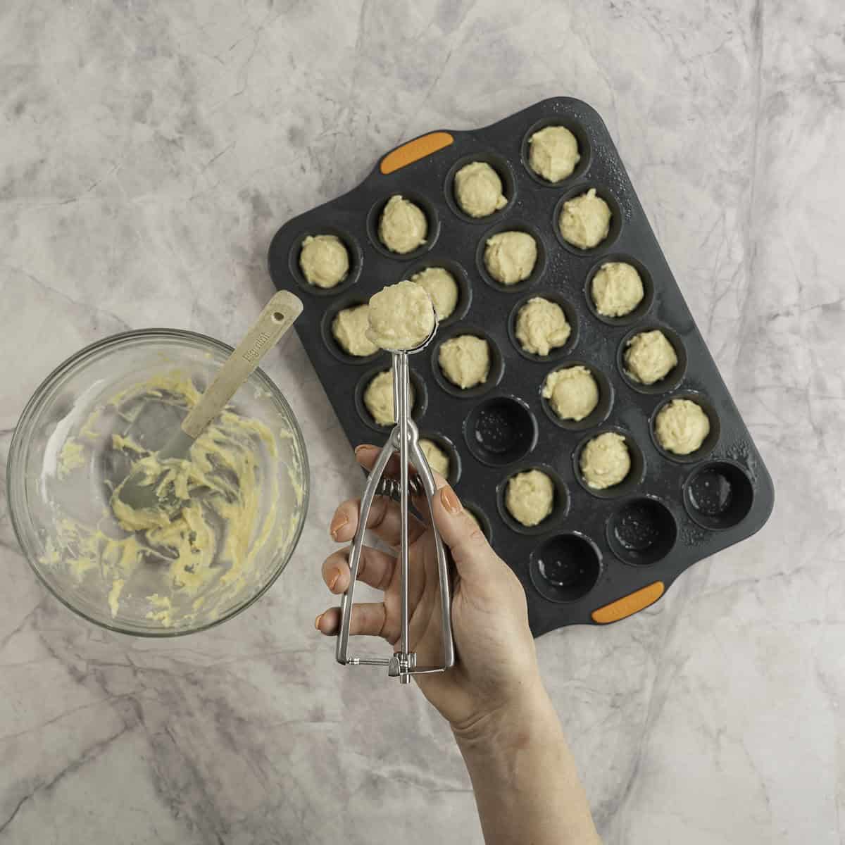 Mini muffin tray with muffin mixture being portioned inside with a small cookie scooper, glass bowl with excess mixture and spatula inside.