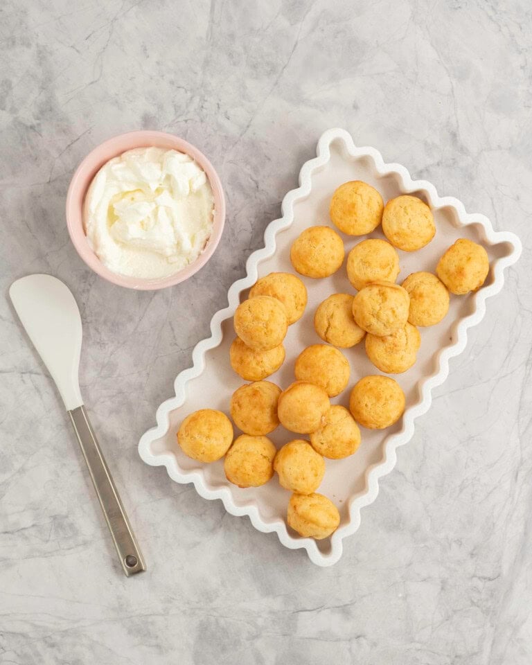 White rectangle dish with wavy border, batch of mini muffins inside, small pink ramekin with greek yogurt inside and a spatula on bench top.
