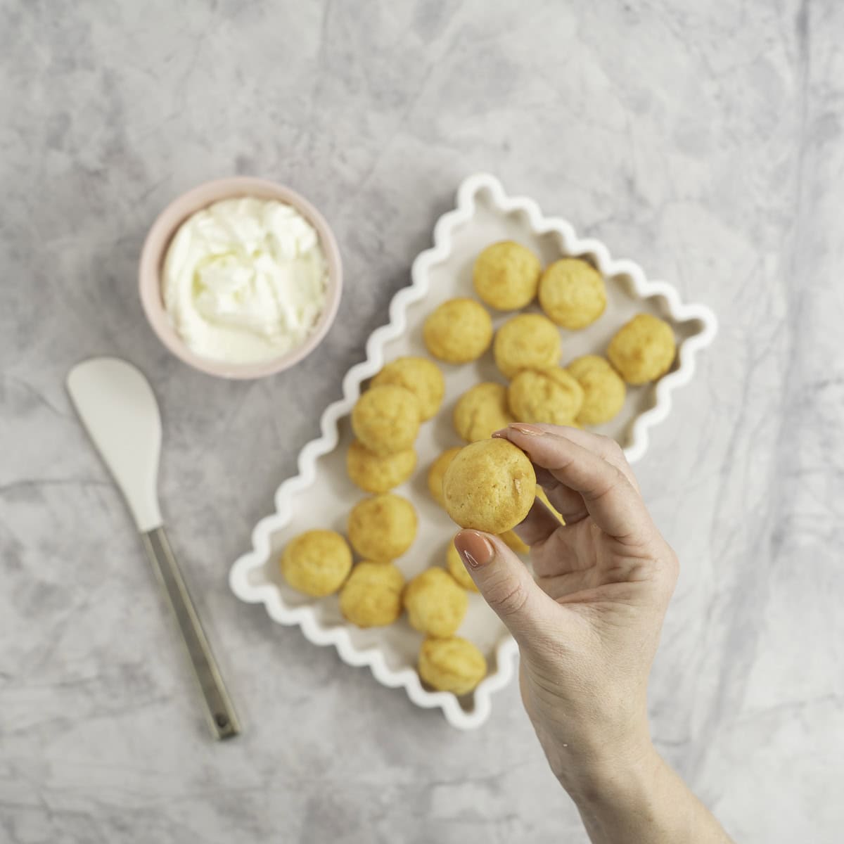 Womans hand holding a mini muffin above a white rectangle dish with wavy border, batch of mini muffins inside, small pink ramekin with greek yogurt inside and a spatula on bench top.
