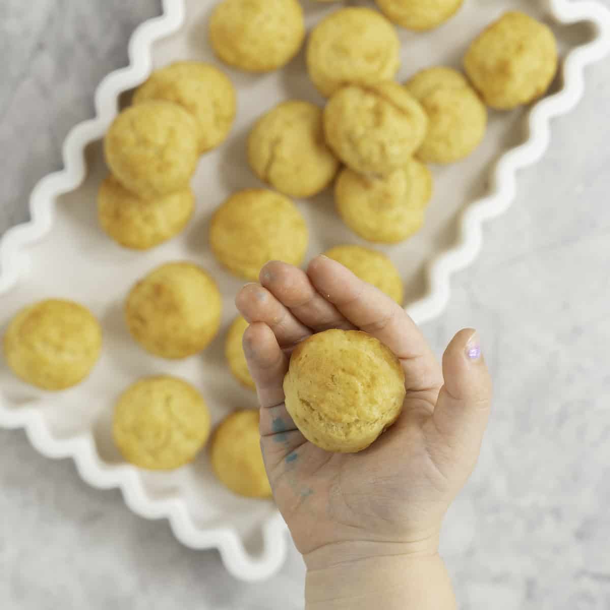 Toddlers hand holding a mini muffin above a white rectangle dish with wavy border, batch of mini muffins inside.