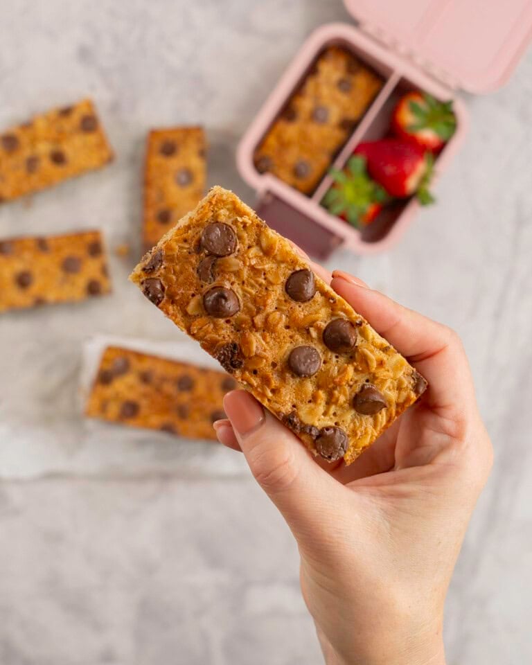 Woman's hand holding a golden granola bar with lunchbox below.