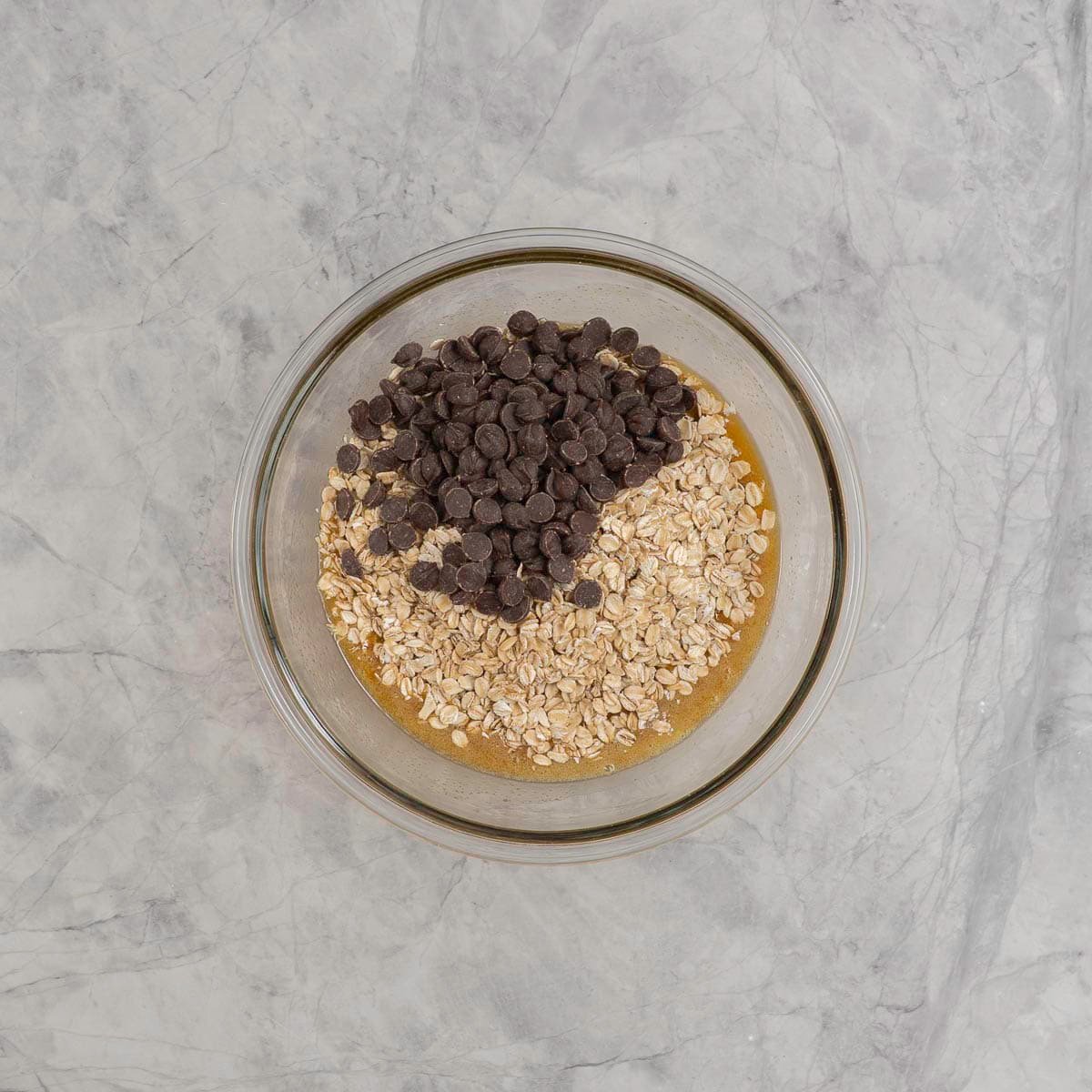 Oats and chocolate chips added into butter mixture in glass bowl on marble benchtop.