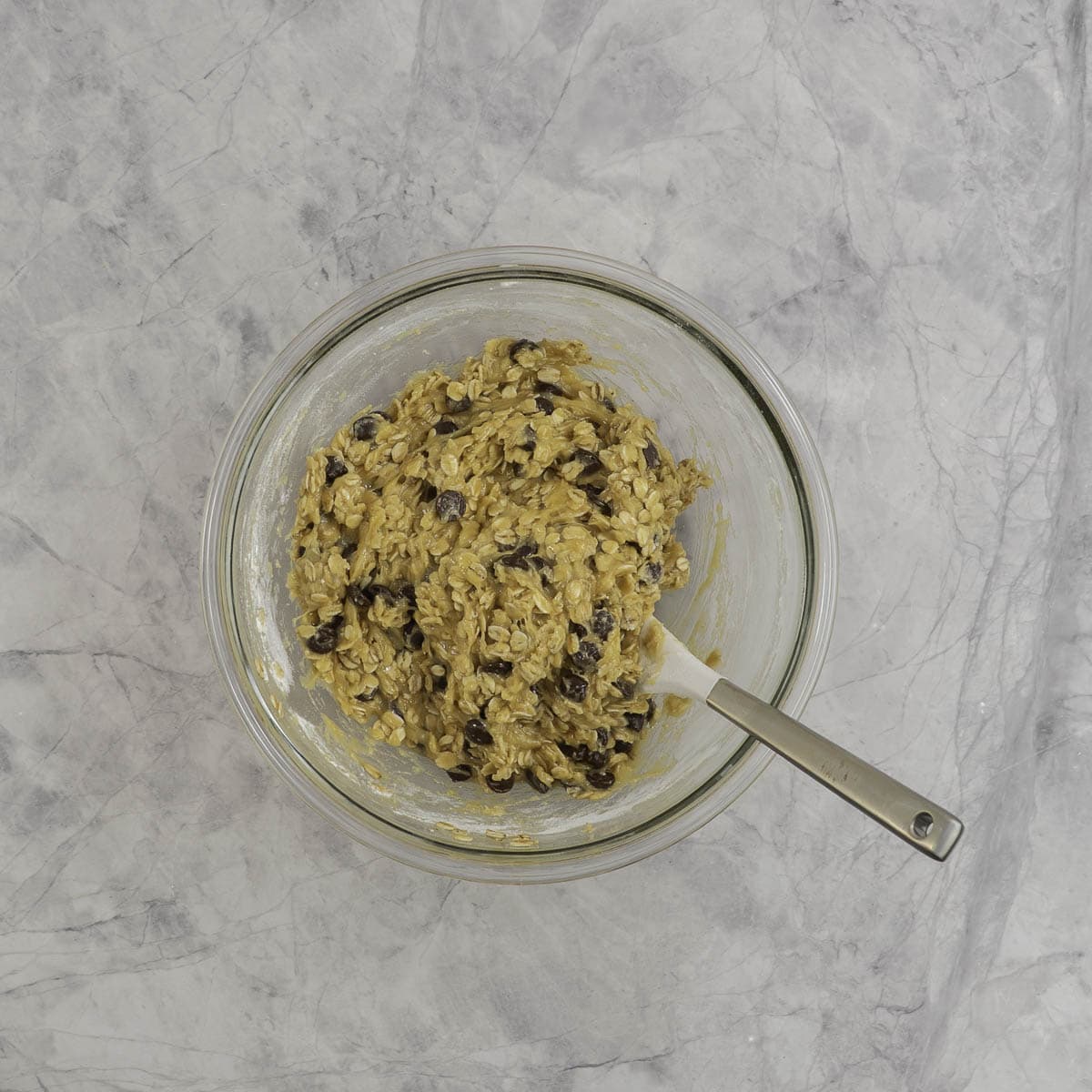 Oats and chocolate chips mixed into butter mixture in glass bowl on marble benchtop.