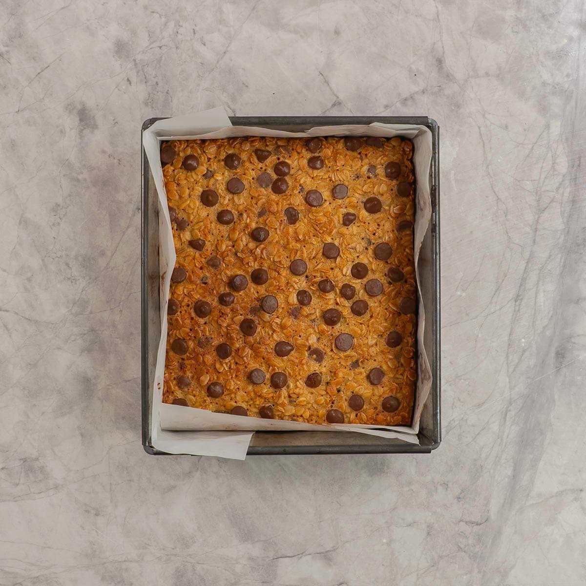 Golden granola bar mixture in a square lined tin with chocolate chips on top.