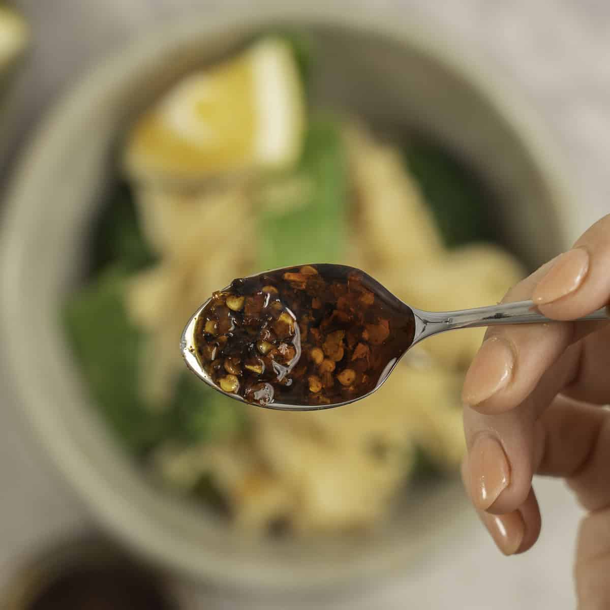 Womans hand holding a teaspoon of chilli sauce above a bowl of peanut butter noodles.