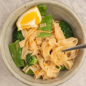 Dinner bowl on benchtop with rice noodles with peanut butter mixture added throughout, broccoli and snow heads inside and garnished with slice of lemon.