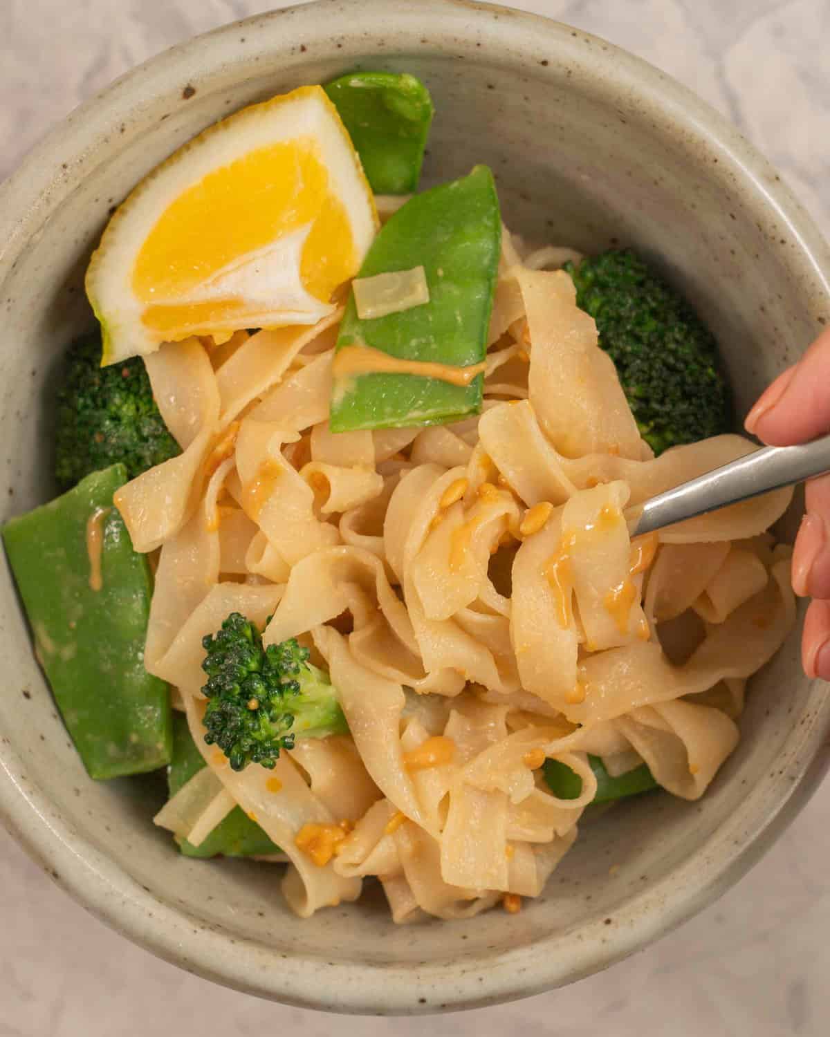 Dinner bowl on benchtop with rice noodles with peanut butter mixture added throughout, broccoli and snow heads inside and garnished with slice of lemon.