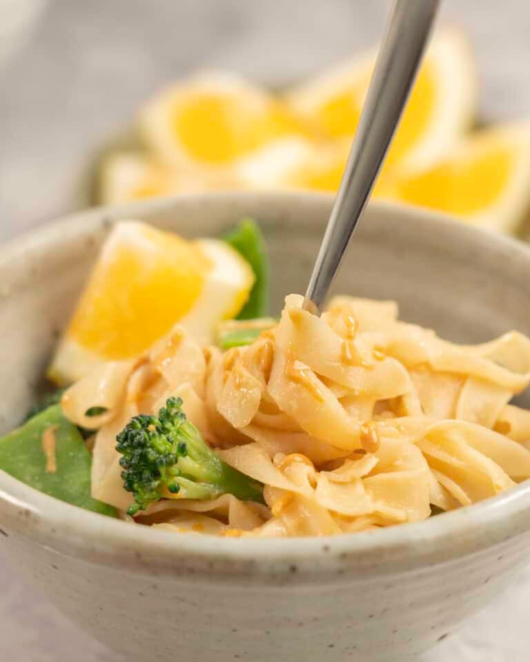Dinner bowl on benchtop with rice noodles with peanut butter mixture added throughout, broccoli and snow heads inside and garnished with slice of lemon.