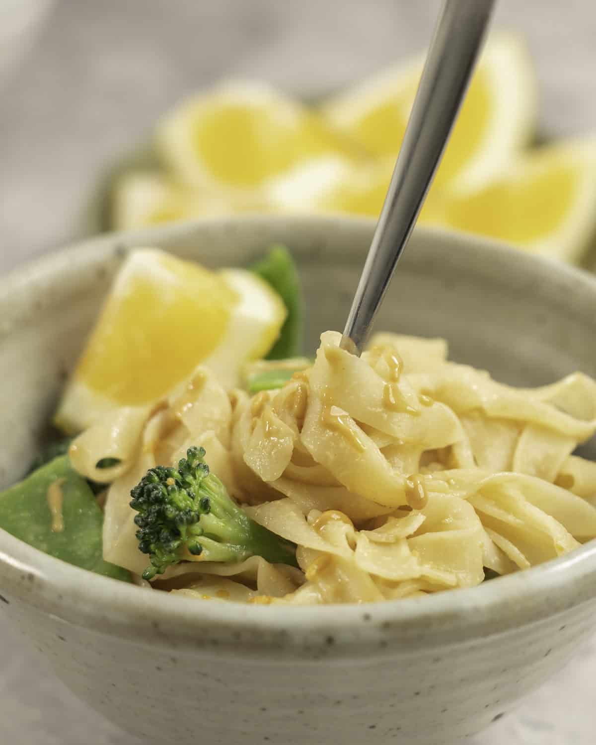 Dinner bowl on benchtop with rice noodles with peanut butter mixture added throughout, broccoli and snow heads inside and garnished with slice of lemon.