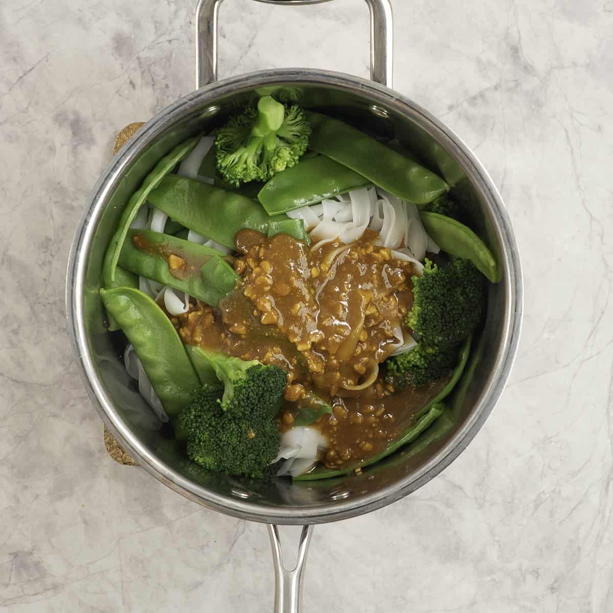 Pot of cooked rice noodles, snow peas and broccoli heads with peanut sauce mixture added on top.