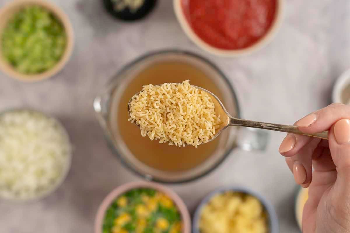 Woman's hand holding a desert spoon with uncooked pasta alphabet letters above blurred ingredients in bowls on benchtop below.