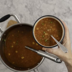 Woman's hand holding a small bowl with spoon inside and vegetable soup with alphabet pasta, pot of soup below on benchtop with a ladle.
