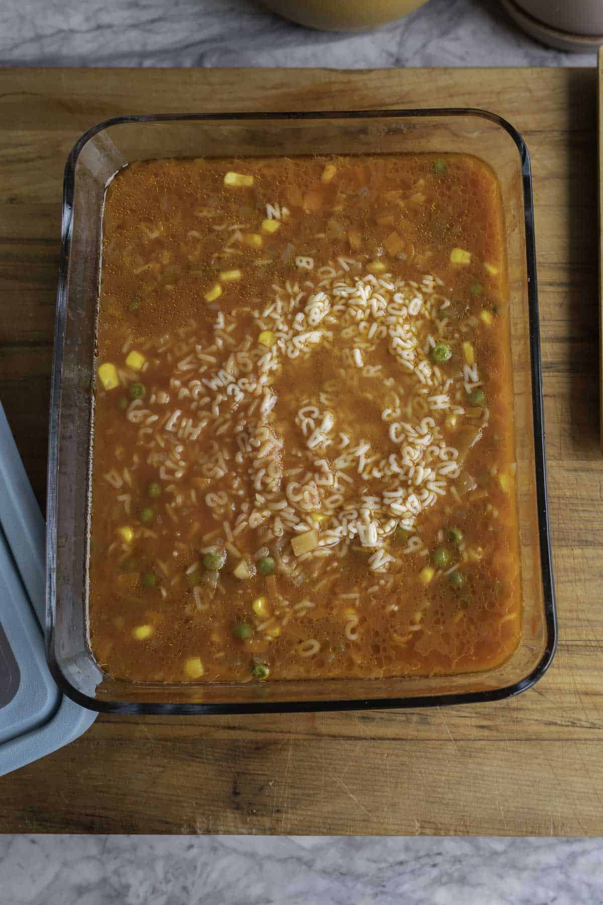 Large glass rectangle container on wooden chopping board with cooked soup and alphabet pasta inside.