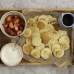 Womans hands holding a tray with mini pancakes on baking paper, a small jug of maple syrup, bowl of yogurt and cut strawberries.