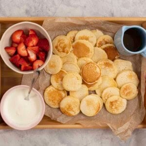 Womans hands holding a tray with mini pancakes on baking paper, a small jug of maple syrup, bowl of yogurt and cut strawberries.