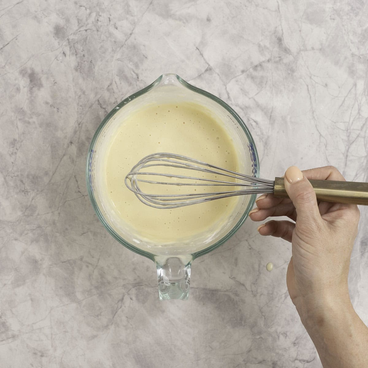 Woman holding a whisk above a glass jug of pancake mixture.