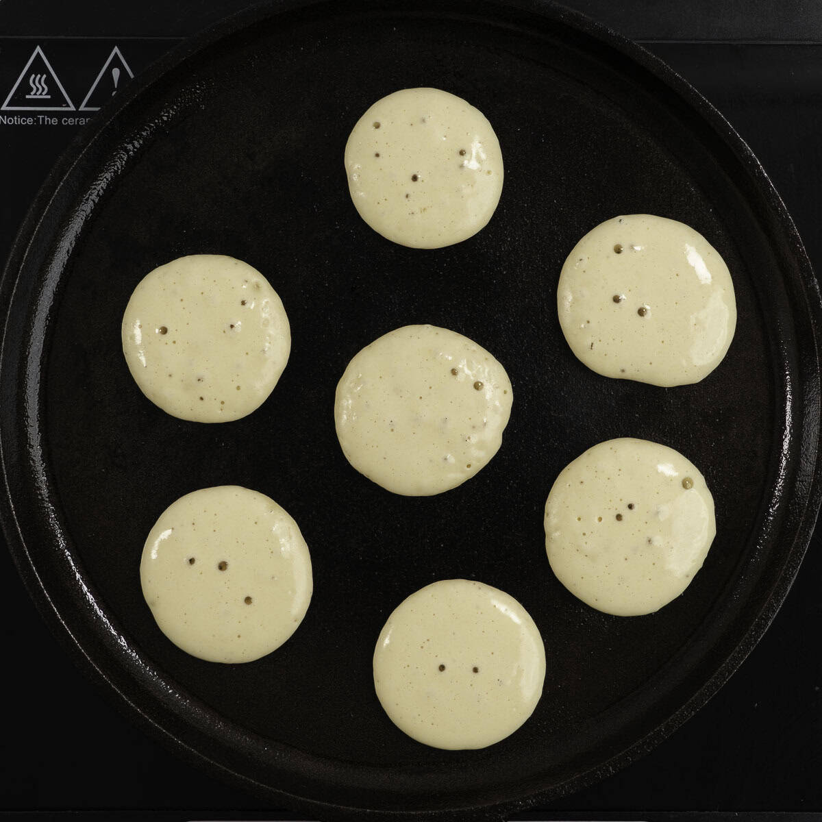 Seven small portions of pancake mixture in a fry pan with bubbles on the top.