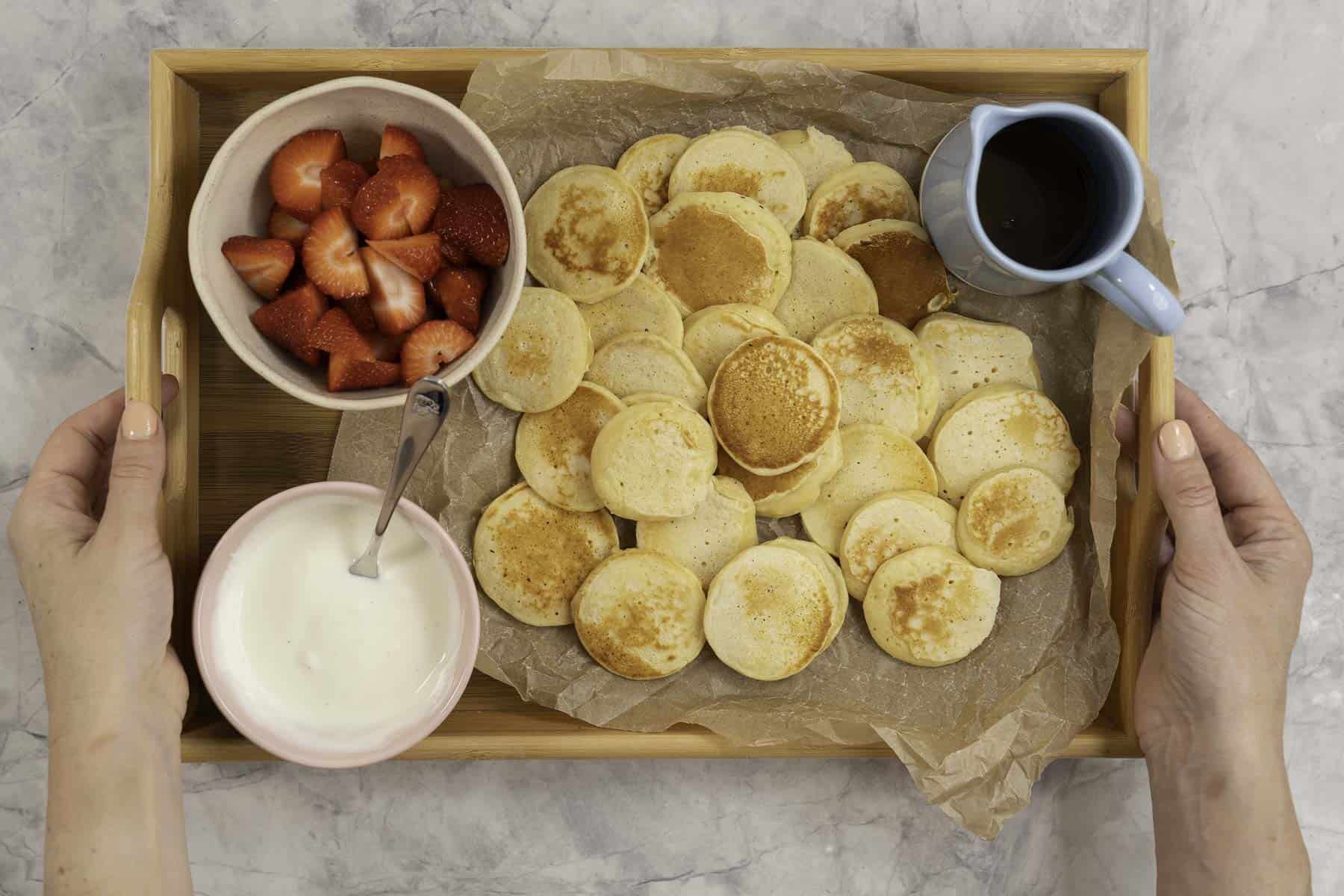 Womans hands holding a mini pancake, below on a tray lined with baking paper a spread of pancakes, a small jug of maple syrup, bowl of yogurt and cut strawberries.