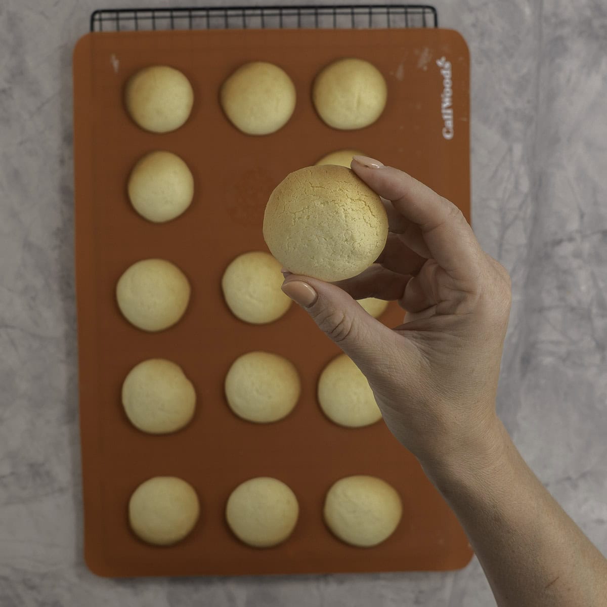 Womans hand holding up to camera a small cookie with more mini cookies on silicone tray below.
