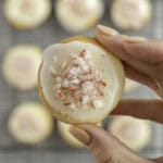 Woman's hand holding a small cookie with icing and crushed candy cane pieces on top, above a cooling tray with more cookies below.