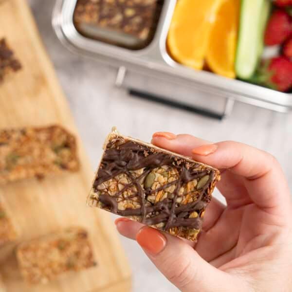 Womans hand holding a small square granola bar with drizzled melted chocolate.