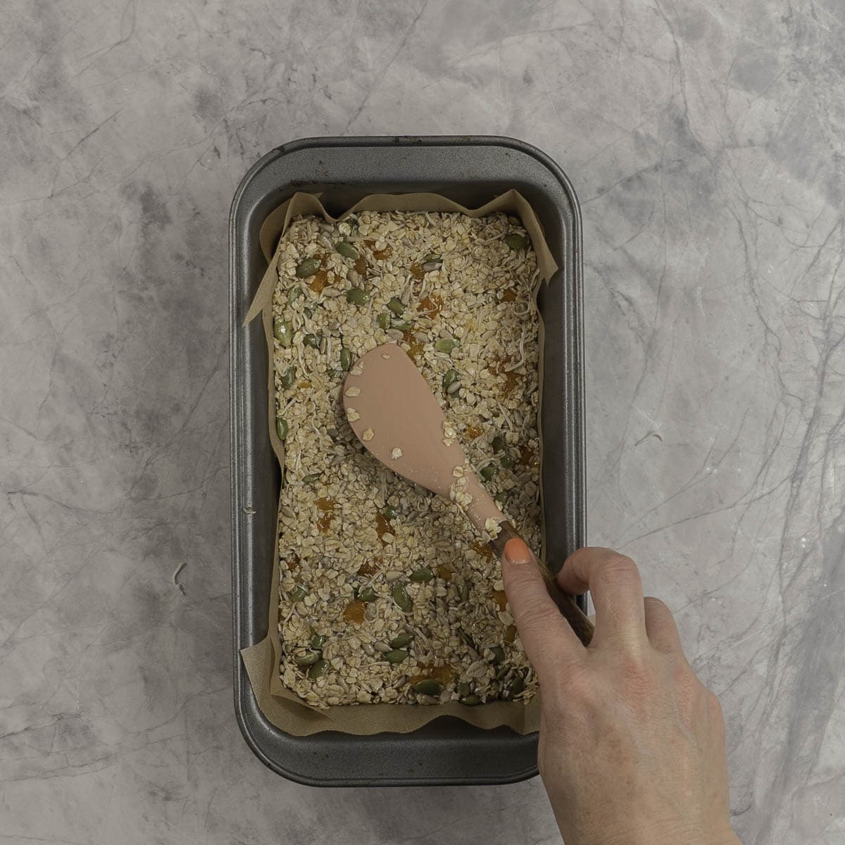 Womans hand holding a spatula pushing nut free bar mixture into a lined rectangle baking tray.