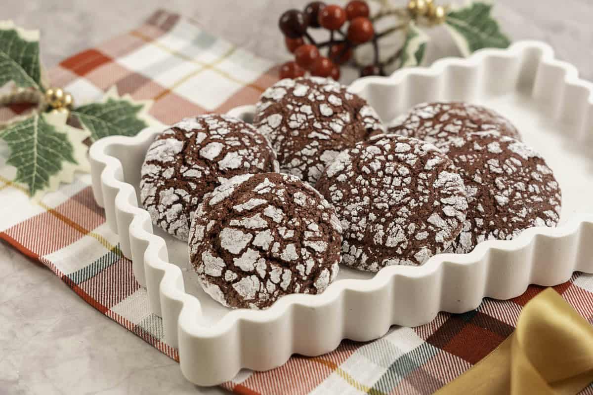 Red coloured cookies with cracked outta shell, coated in icing sugar placed in a white dish on christmas decorations in background and red and green checkered tea towel below.